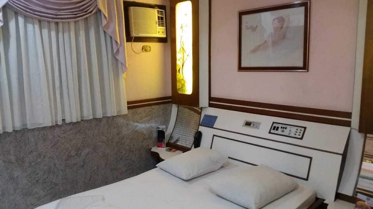 Ein Hotelzimmer mit Bett, Nachttisch mit Lampe, Fenster mit Vorhängen, Klimaanlage und einem gerahmten Bild an der Wand.