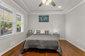 Une chambre à coucher comprenant un lit, un ventilateur de plafond, une fenêtre avec des stores, une table de chevet et une œuvre d’art accrochée au mur.