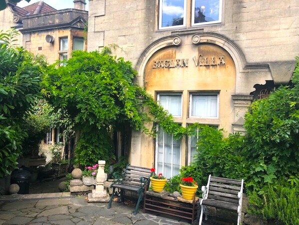 A building with a sign reading "RACHAU VILLA". There are two wooden benches in front of the building, surrounded by greenery and potted plants.