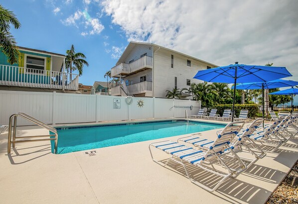 A swimming pool with lounge chairs and umbrellas, a white building with balconies, and palm trees.