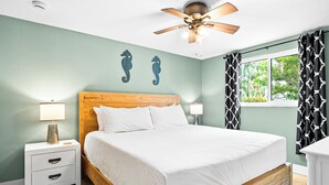 Une chambre à coucher avec un lit en bois, deux tables de chevet, un ventilateur de plafond et une fenêtre ornée de rideaux à motifs.