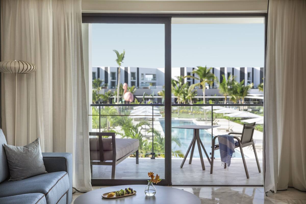 A modern hotel room with a large sliding glass door leading to a pool area, a sofa, a coffee table with a vase, and a view of the pool and resort buildings.