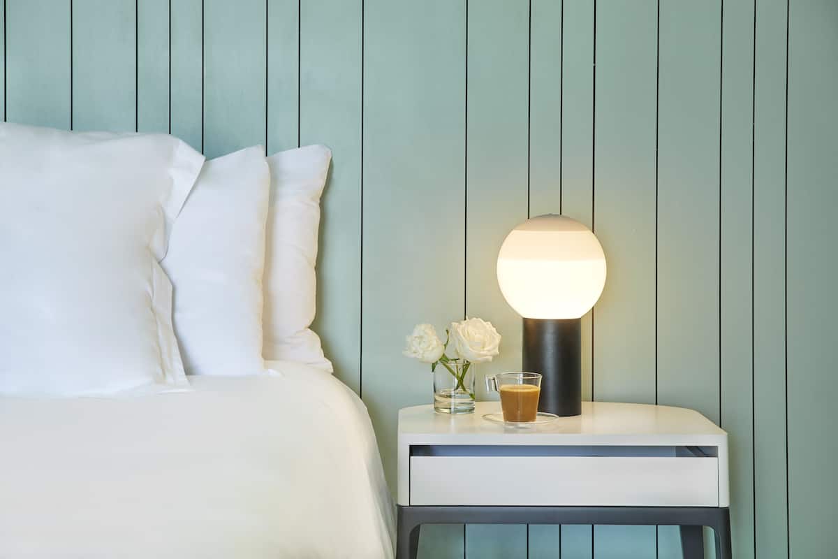 A neatly made bed with white linens and pillows, a bedside table with a lamp, a glass of coffee, and two white roses.