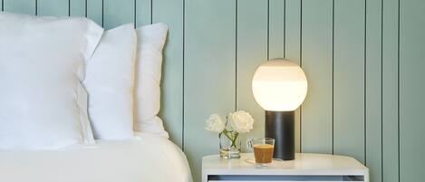 A neatly made bed with white linens and pillows, a bedside table with a lamp, a glass of coffee, and two white roses.