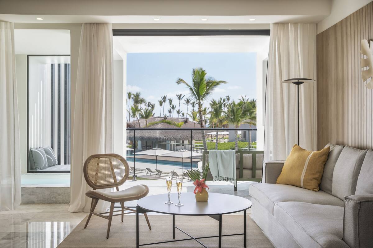 A modern living room with a view of a pool and palm trees, a round coffee table with glasses, and a sofa with cushions.