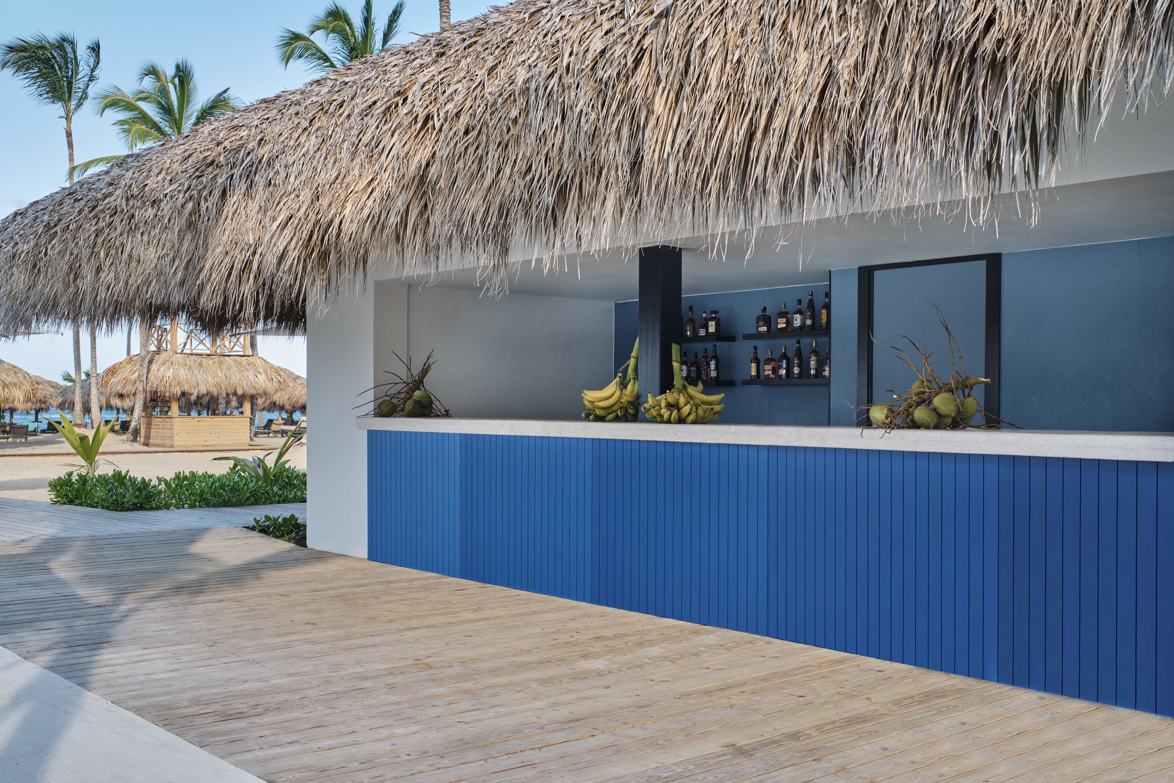 A beachside bar with a thatched roof, blue paneling, and a wooden deck.