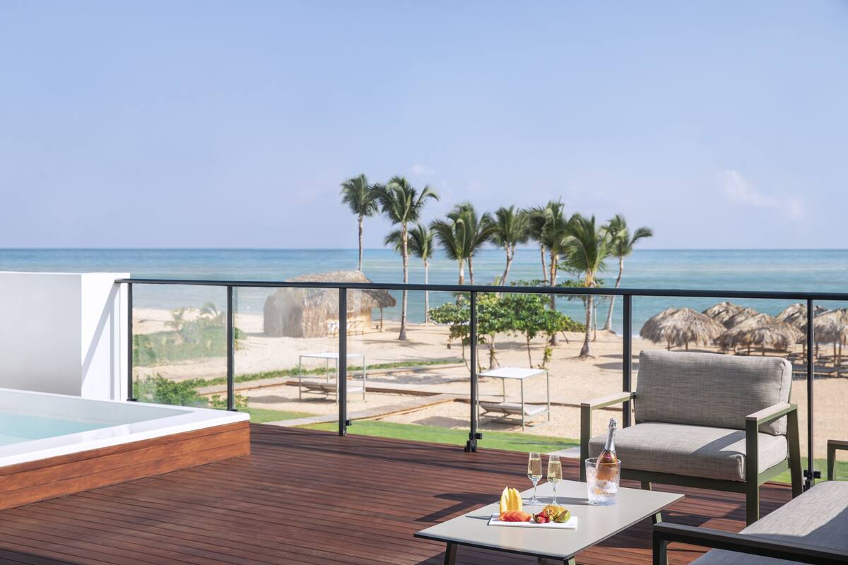 A wooden deck with a pool, a table with fruit and drinks, and a view of the beach and ocean.