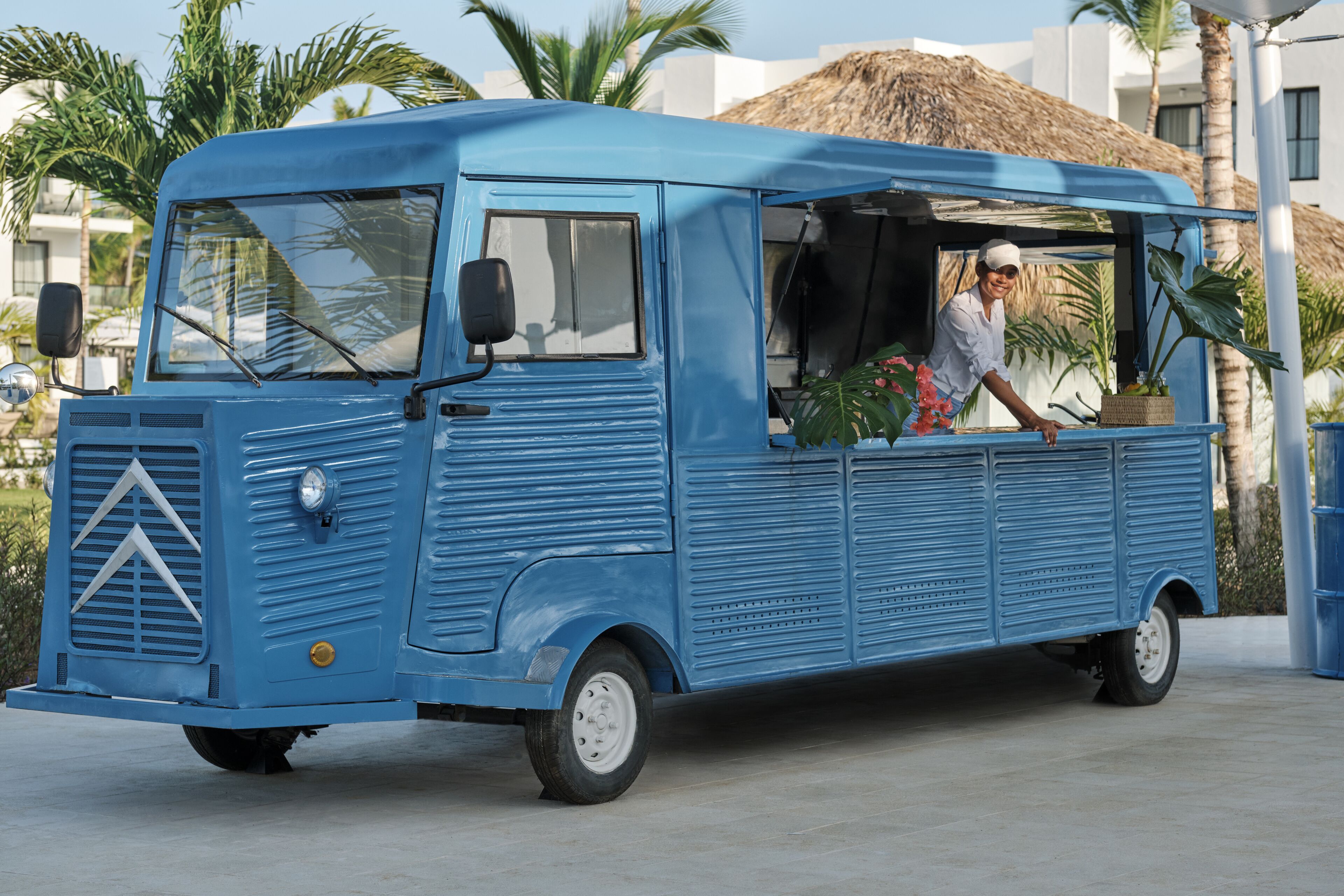 Un vieux fourgon bleu, avec une personne à l’intérieur, est garé dans un espace extérieur ensoleillé, bordé de palmiers, avec un bâtiment en arrière-plan.