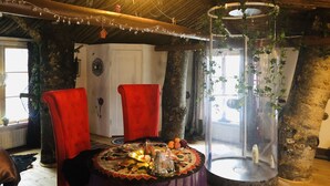 A rustic room with a high ceiling, exposed wooden beams, a round table with a floral tablecloth, two red chairs, and a glass enclosure with plants. - Treehouse Magpies Nest With Bubble Pool (Avesta)