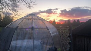 A transparent greenhouse with a table and chairs inside, surrounded by a wooden fence and trees. - Treehouse Magpies Nest With Bubble Pool (Avesta)