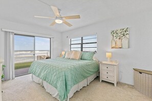 Una camera da letto con un letto, un comodino, una lampada, una ventola a soffitto, una finestra con persiane e vista sull'oceano.