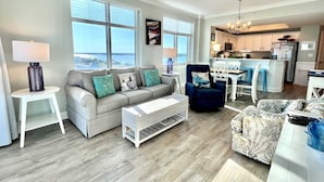 Un salón con un sofá gris, un sillón azul y un sillón estampado. Hay una mesa de centro blanca y una mesita auxiliar blanca con una lámpara. La habitación tiene ventanas amplias con vista al océano.