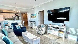 Una sala moderna con un televisor de pantalla plana grande, un conjunto de sofás, una mesa de centro y un comedor con una cocina al fondo.
