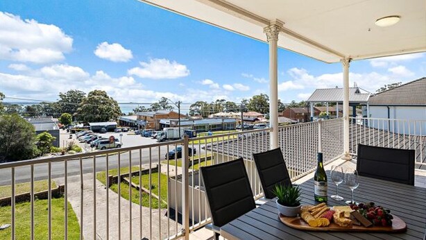 A balcony with a view of a parking lot and buildings, a table with a bottle of wine, and a tray of snacks. - Vincentia Breeze by Experience Jervis Bay (Vincentia)