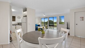 A modern kitchen with white cabinets, a refrigerator, and a dining area with a round table and white chairs. - Ocean Outlook 2 on Marine Parade (KINGSCLIFF)
