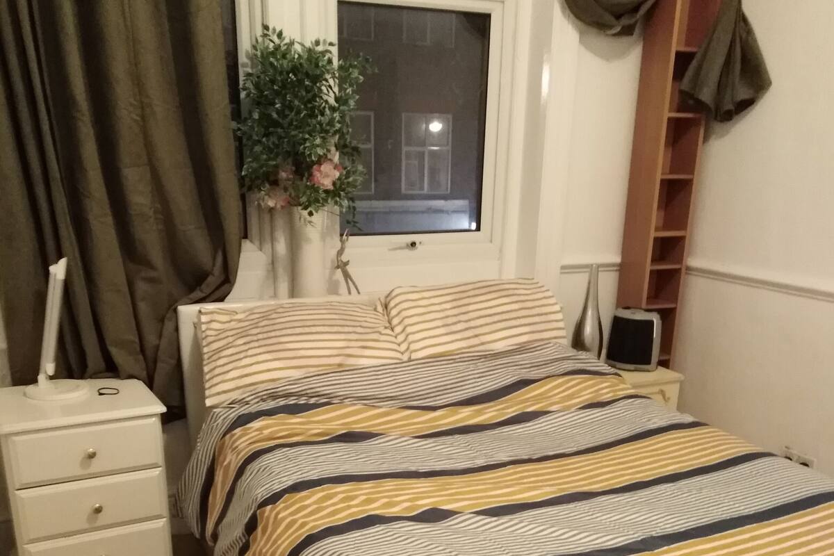 A bedroom with a bed, a nightstand, a dresser, a window with curtains, a bedside table, a lamp, a vase with flowers, and a shelf.