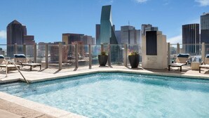En pool på taket med solstolar och krukväxter, med utsikt över stadens silhuett och moderna byggnader.