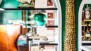 Una habitación con una pared verde, un sillón de cuero, una mesa de vidrio con libros, una columna decorativa dorada y un espejo con marco blanco.