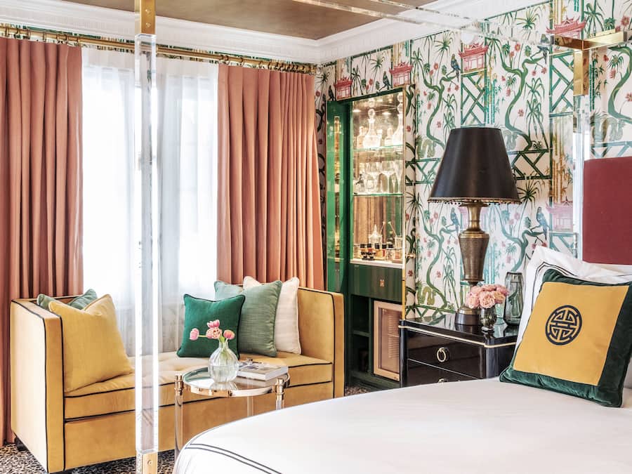 A hotel room with a bed, two armchairs, a small table with a vase, a lamp, and a nightstand with a mirror and decorative items.