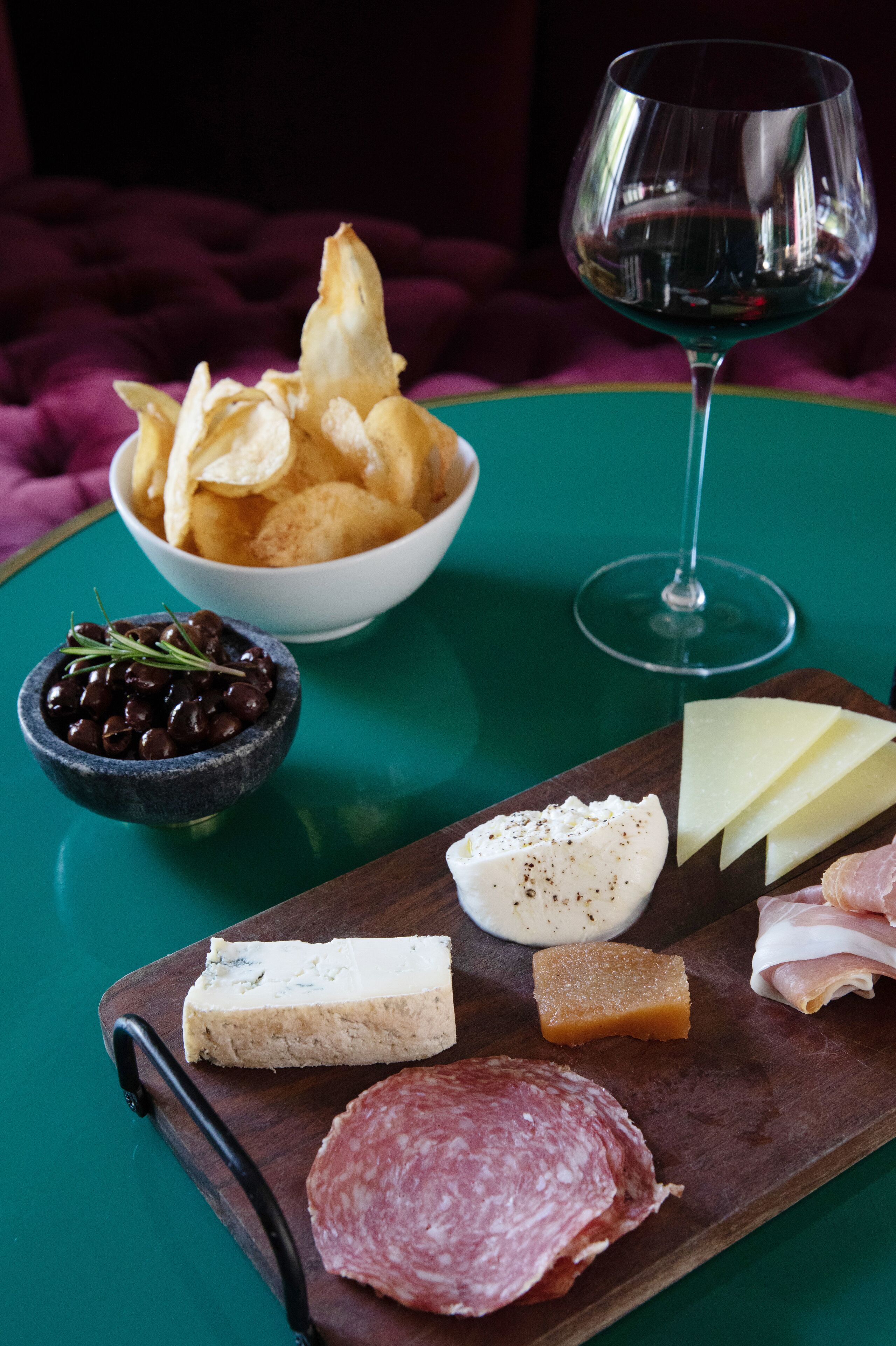 A charcuterie board with various cheeses, cured meats, olives, and crackers on a wooden board, accompanied by a glass of wine on a green table.