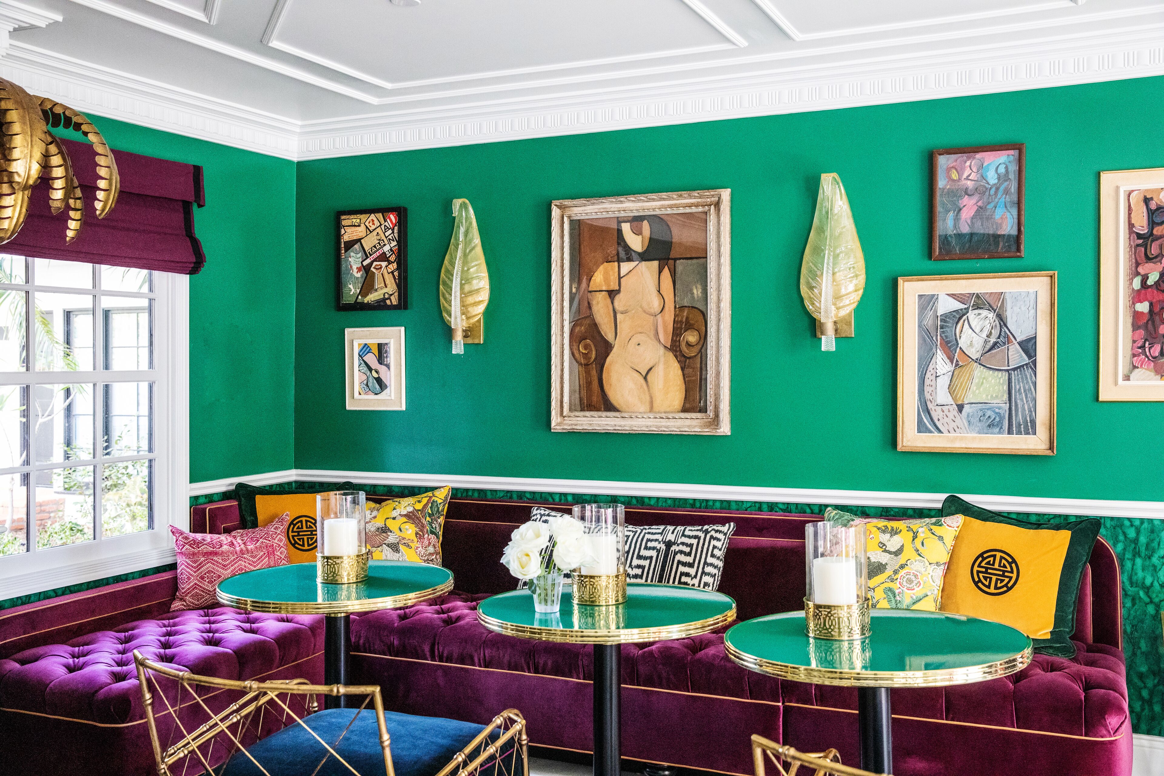 Ein Zimmer mit grünen Wänden, einem grünen Sofa und einem Tisch mit goldenen Akzenten. An den Wänden befinden sich gerahmte Kunstwerke.