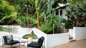 Un patio con dos sillas negras y una pequeña mesa, rodeado de abundante vegetación y una pared blanca con chimenea decorativa.