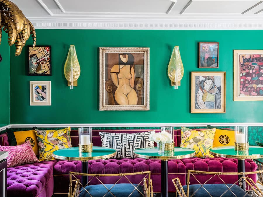 Una habitación vibrante con paredes color turquesa, un sofá morado mullido y varias mesas redondas con detalles dorados.