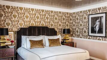 A bedroom with a large bed, ornate wallpaper, a chandelier, and a framed picture on the wall.