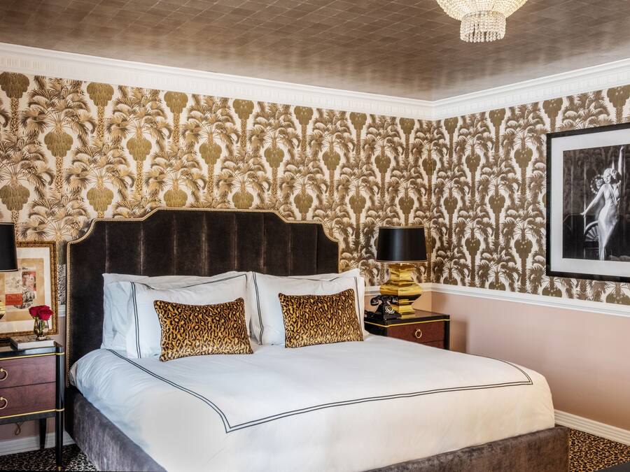 A bedroom with a large bed, ornate wallpaper, a chandelier, and a framed picture on the wall.