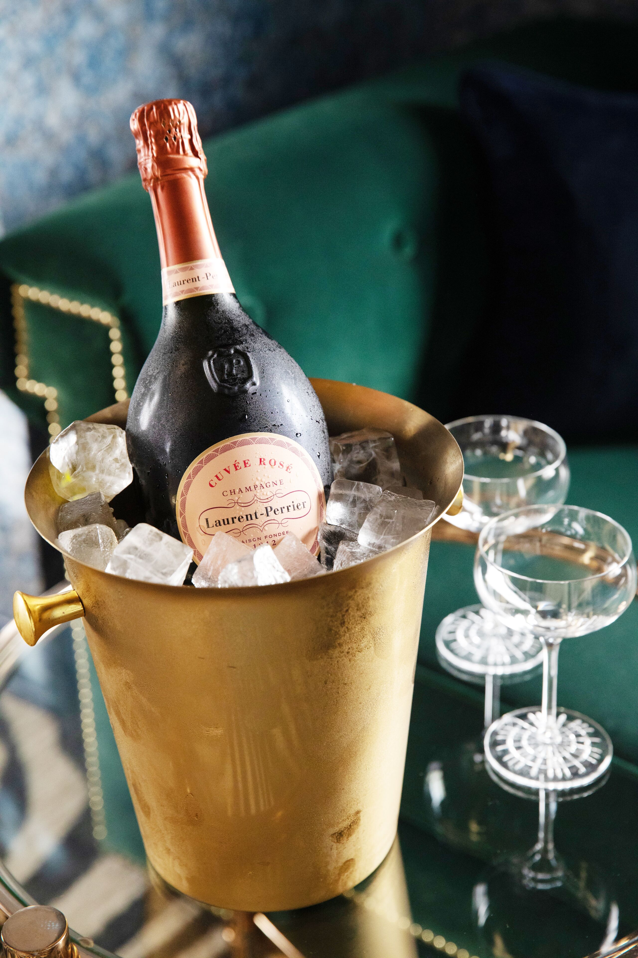 A bottle of champagne in a gold bucket with ice cubes, two empty glasses, and a green upholstered sofa in the background.
