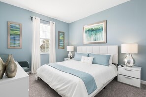 A bedroom with a large bed, two bedside tables, a nightstand, a window with curtains, and two framed artworks on the wall.