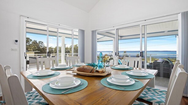 Een eetruimte met een houten tafel voor zes personen, grote ramen met uitzicht op de zee, en een balkon met buitenmeubels.