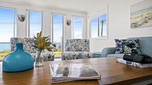 Een woonkamer met een houten salontafel, een tijdschrift en een vaas met bloemen. Er zijn twee gestreept fauteuils en een blauwe bank met een bloemenkussen. Grote ramen bieden uitzicht op de zee.