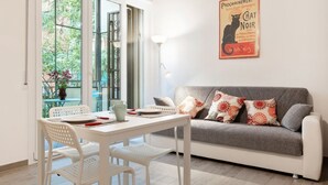 Una sala de estar moderna con un sofá gris, una mesa y sillas blancas, una ventana con vistas a vegetación y un póster en la pared.