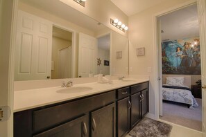 A bathroom with a double sink vanity, a large mirror with lights, and a door leading to a bedroom with a bed and a painting. - 9009 Solara (Kissimmee)