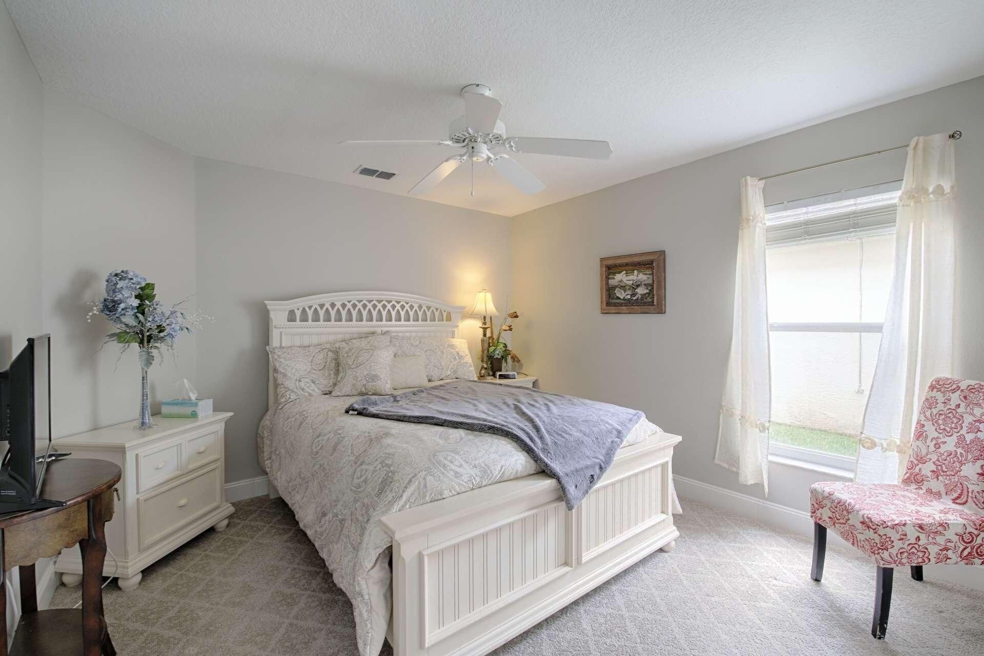 A bedroom with a white bed, a desk, a chair, a ceiling fan, and a window with curtains.