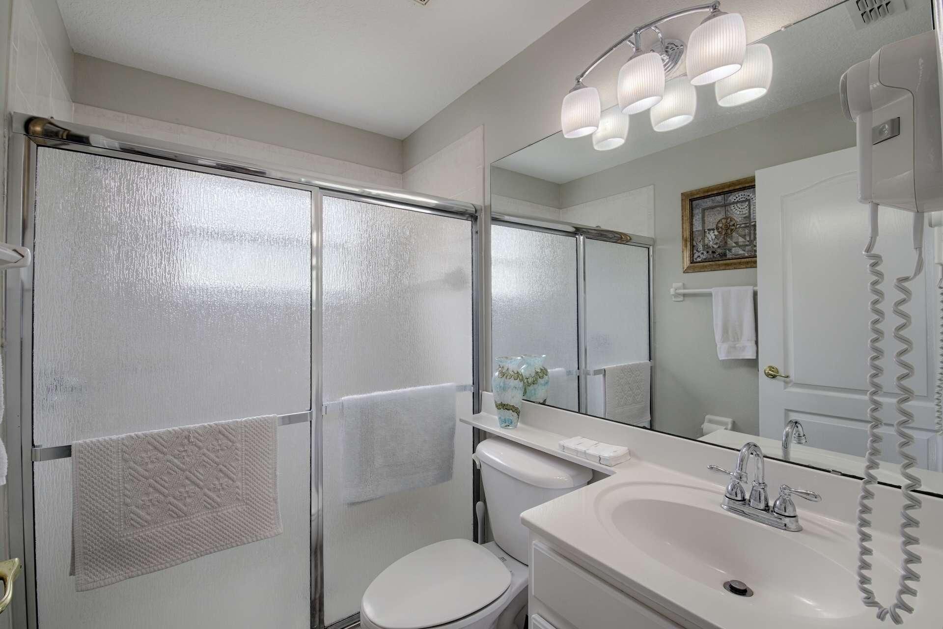 A bathroom with a glass shower enclosure, a toilet, a sink with a mirror, and a telephone.
