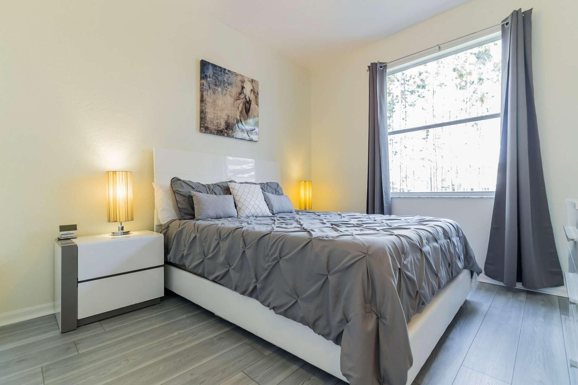 A bedroom with a bed, two bedside tables, a nightstand, a painting, a window with curtains, and a view of trees.