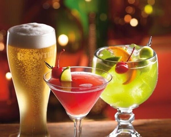 Tres tipos diferentes de bebidas en vasos, incluyendo una cerveza, un cóctel rojo y un cóctel verde, cada uno adornado con frutas y una cereza.
