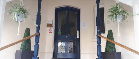 A hotel entrance with a dark blue door, numbered 31, flanked by two potted plants and a wooden handrail.