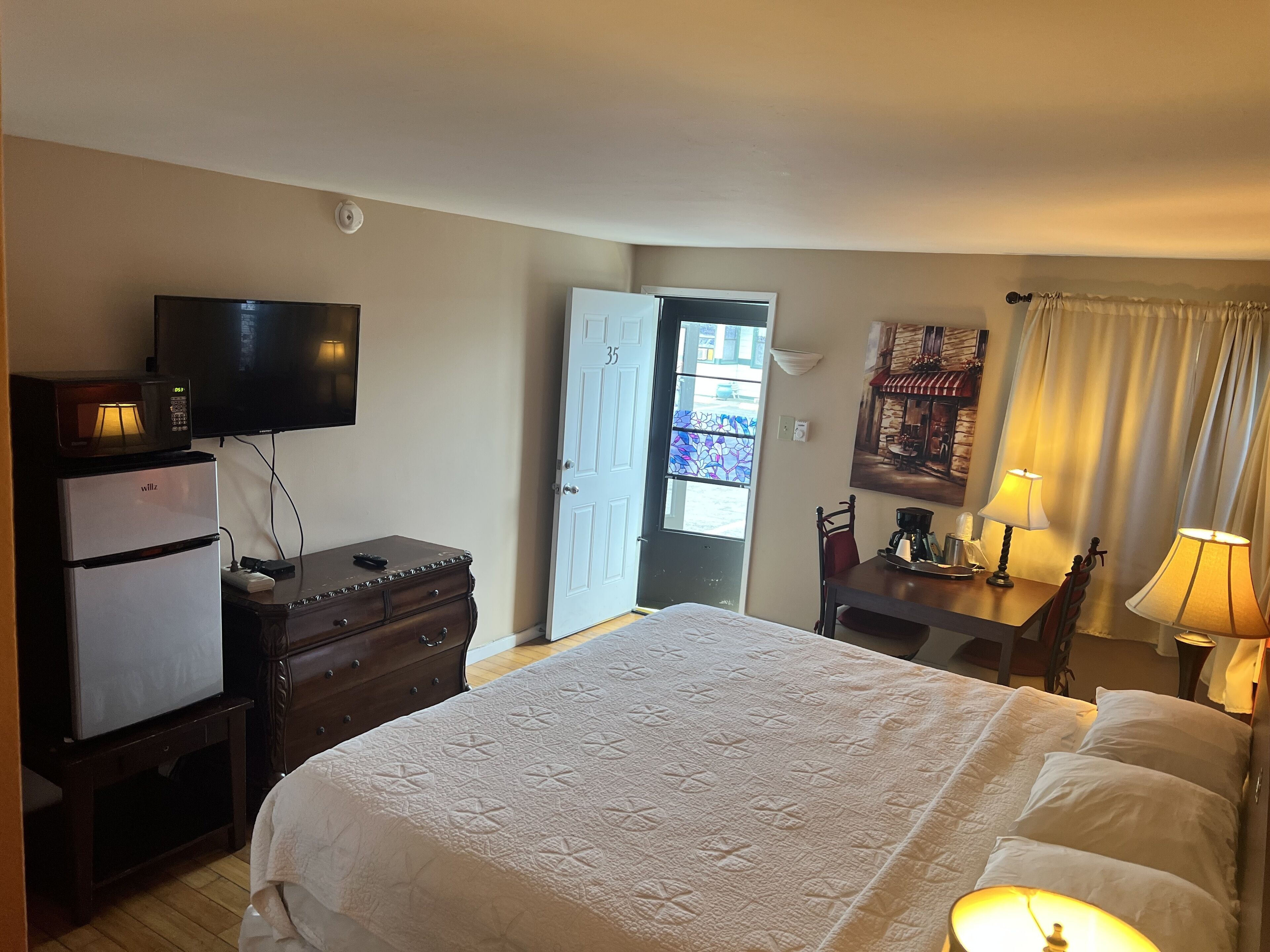 A hotel room with a bed, a television, a refrigerator, a dresser, a desk with a lamp, and a door leading to a balcony.