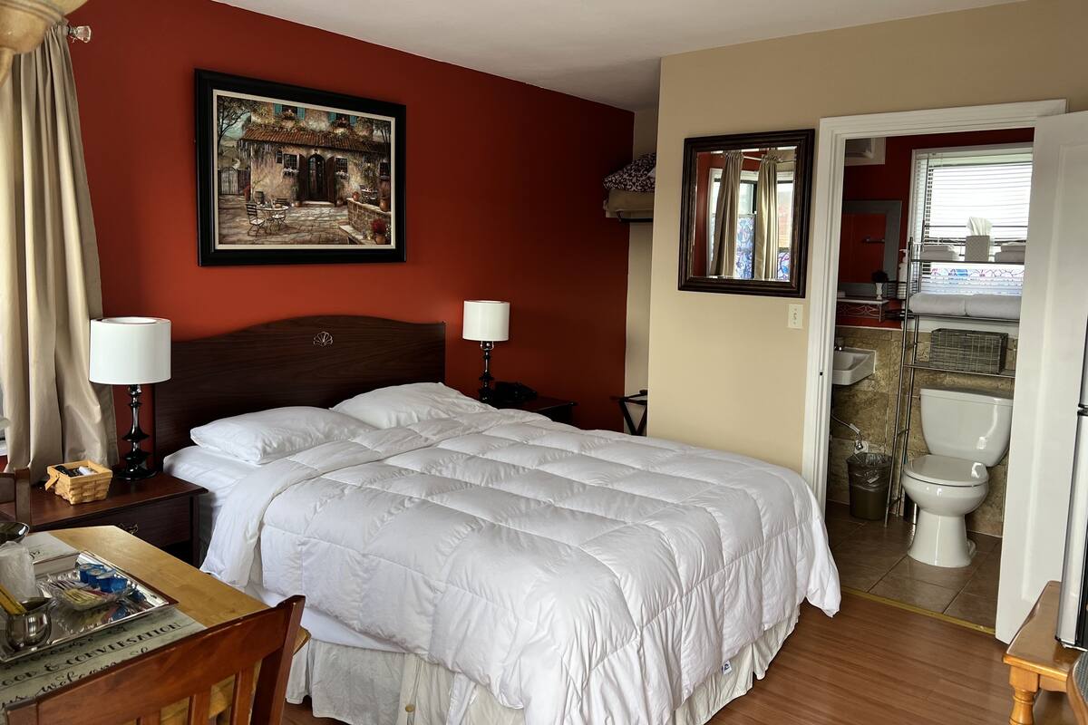 Quarto de hotel com cama, luminárias de cabeceira, escrivaninha com abajur, um quadro na parede e vista para o banheiro.