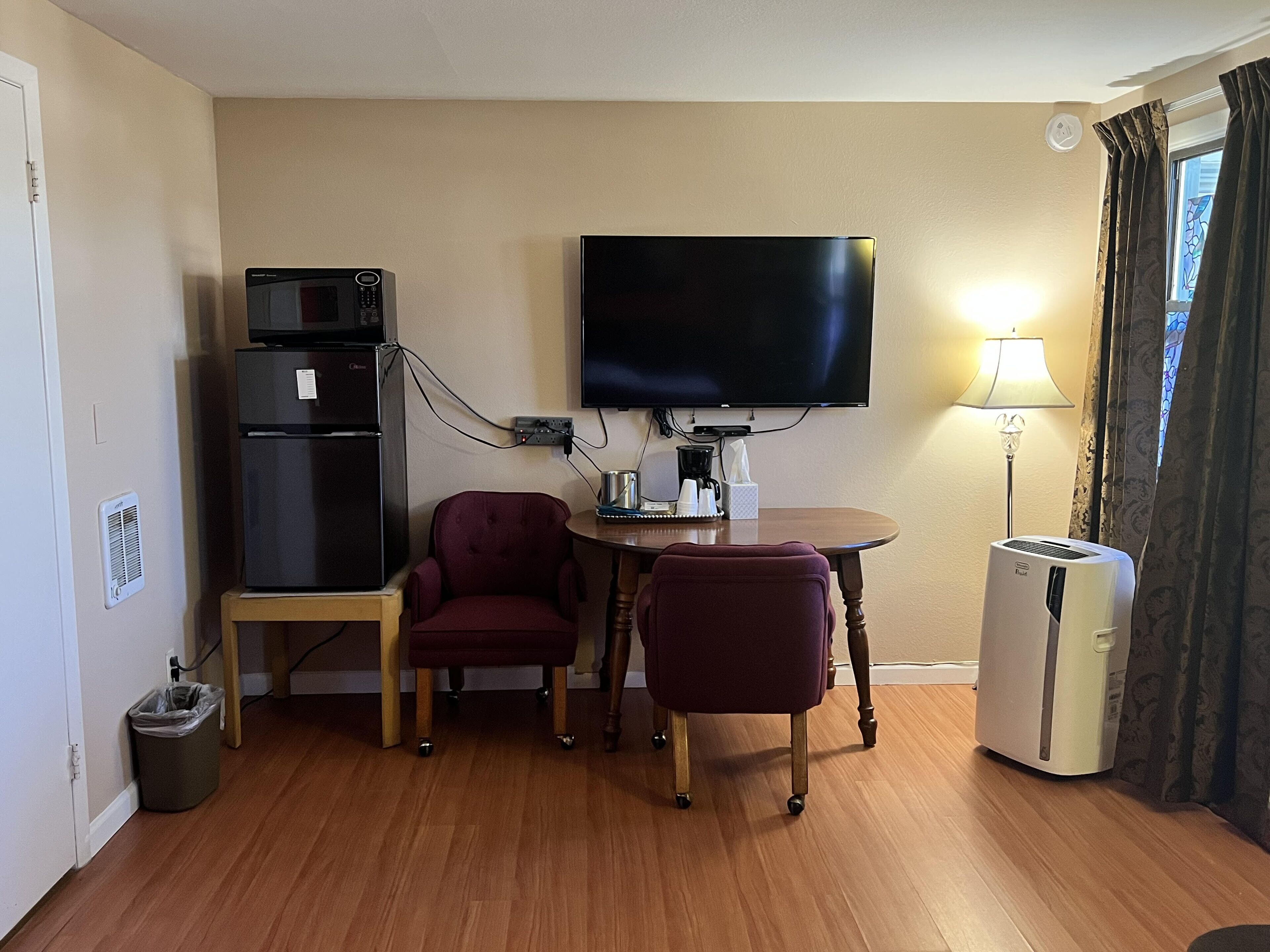Une chambre d’hôtel compacte équipée d’une télévision à écran plat, d’un petit réfrigérateur, d’une table avec deux chaises, d’un radiateur et d’une fenêtre avec des rideaux.