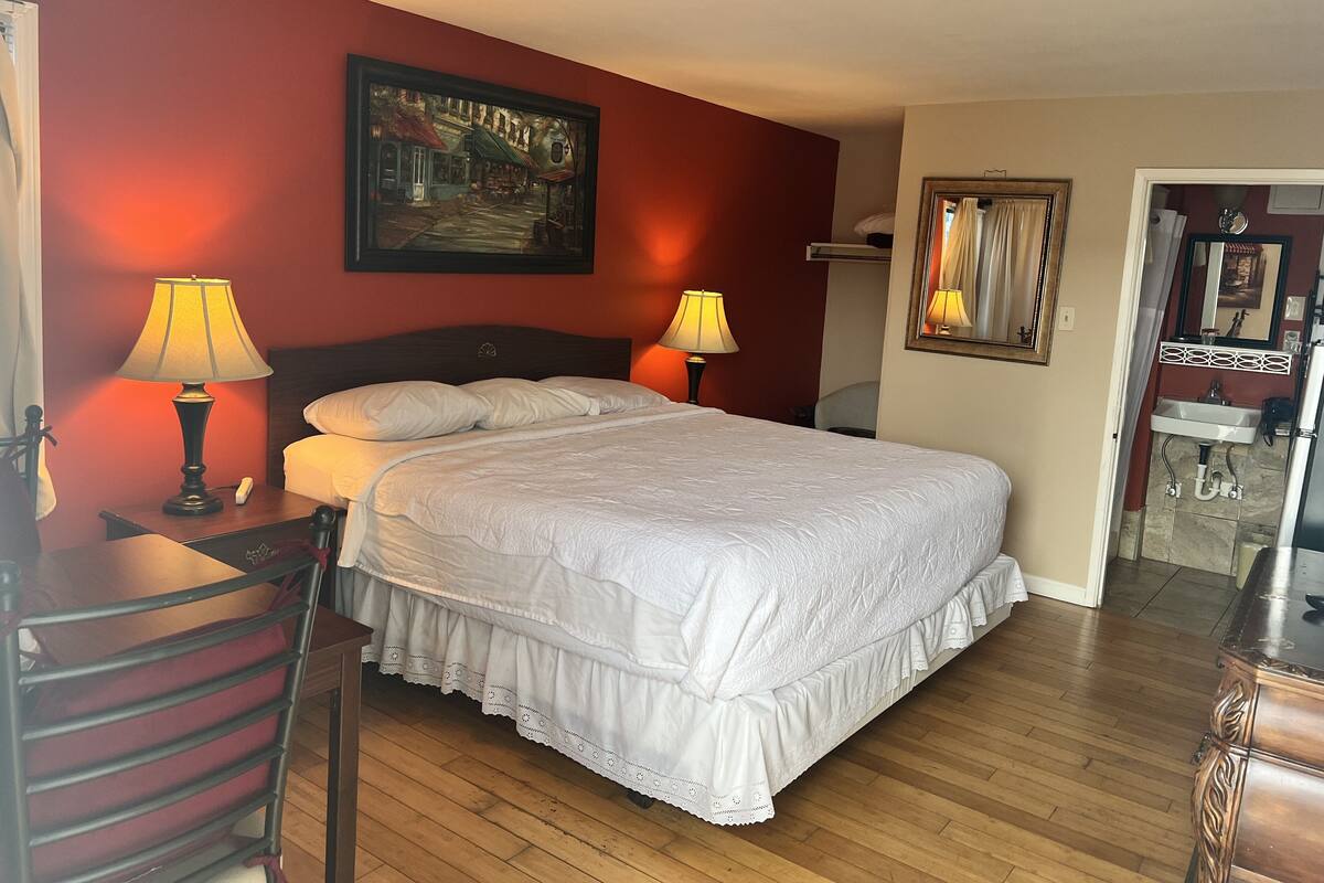 Quarto de hotel com uma cama grande, duas luminárias de cabeceira, um quadro na parede, uma cômoda de madeira e um banheiro visível através de uma porta aberta.