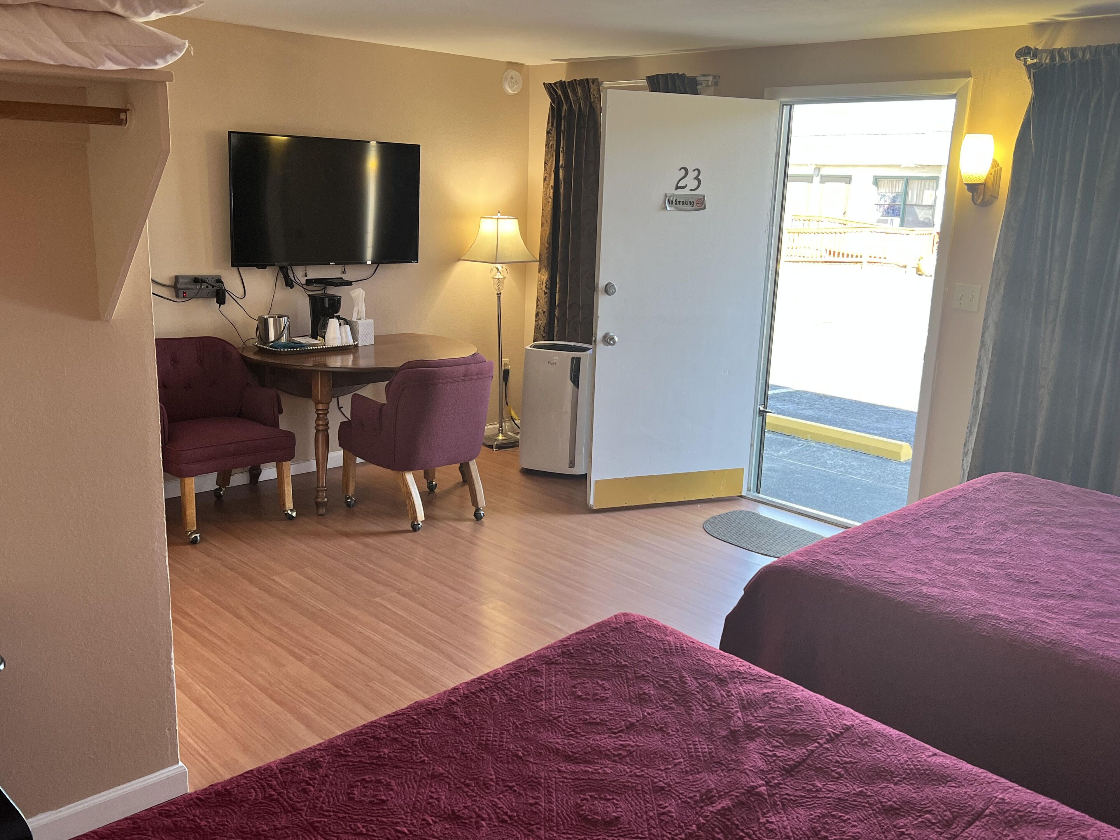 A hotel room with two beds, a desk with a chair, a television, a door with a 'No Smoking' sign, and a view of the outside.