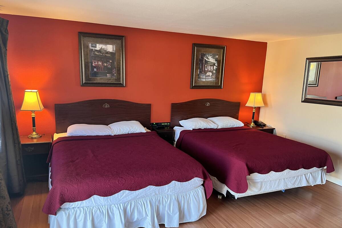 Quarto de hotel com duas camas, cada uma com um cobertor vermelho e almofadas brancas. Há duas imagens emolduradas na parede, uma luminária a cada lado das camas, e piso de madeira.