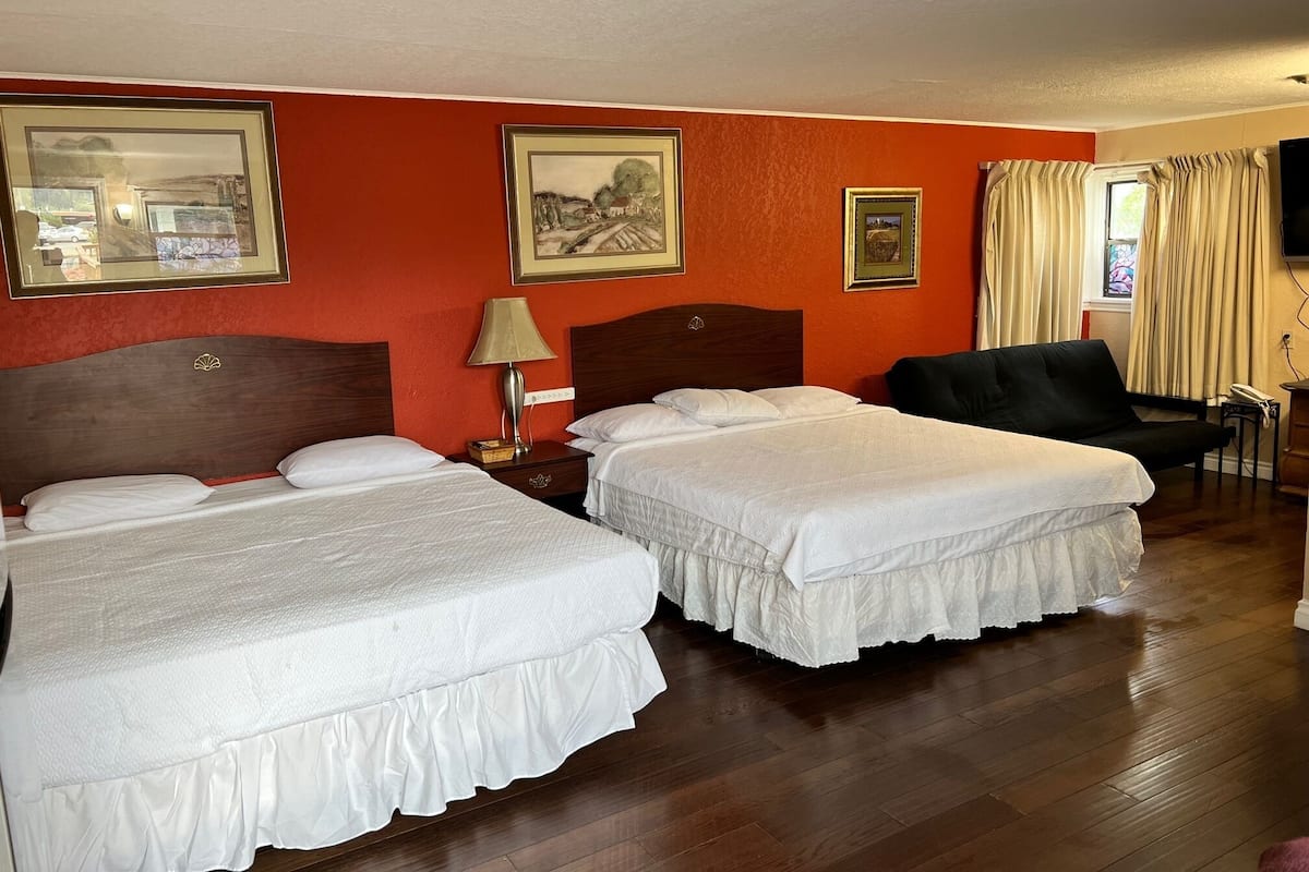 Quarto de hotel com duas camas, uma cabeceira de madeira, mesa de cabeceira com abajur, uma cadeira preta e uma televisão.
