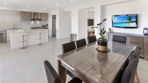 A modern kitchen with white cabinets, a stainless steel sink, and a large island with bar stools. There is a dining area with a wooden table and chairs, and a TV mounted on the wall displaying a tropical beach scene.