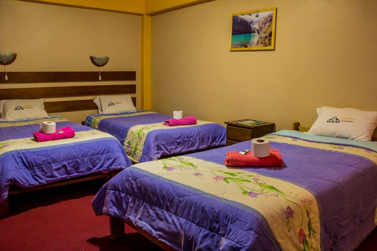 Ein Hotelzimmer mit drei Betten, jedes mit einer violetten Tagesdecke und floralem Muster. Es gibt Kissen mit dem Hotel-Logo, ein Gemälde einer Berglandschaft an der Wand und einen kleinen Tisch mit einer Lampe.