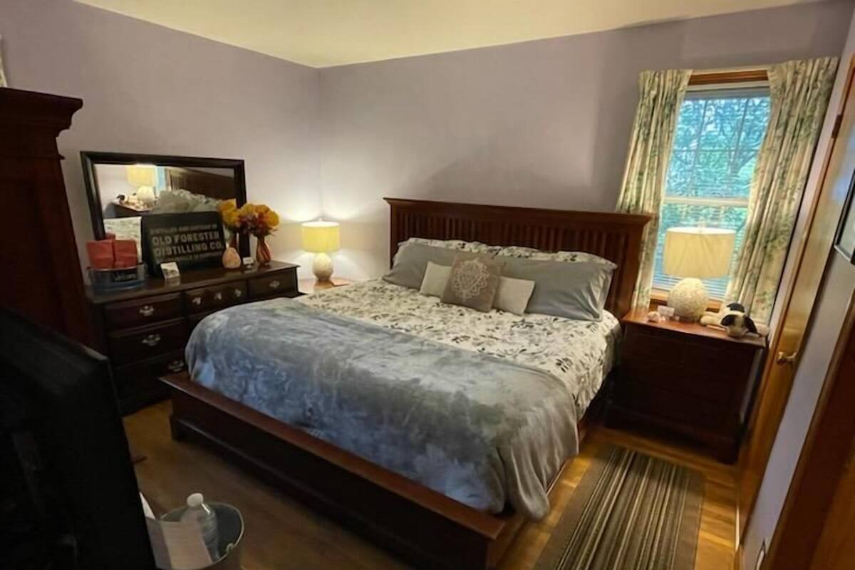 Une chambre à coucher avec un grand lit, une commode, un miroir, une table de chevet et une fenêtre avec des rideaux.